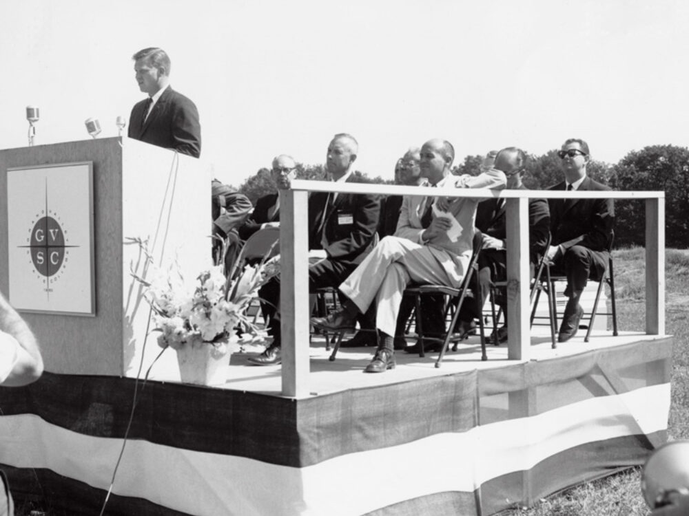 One man speaks at a podium while others are seated on a stage behind him.